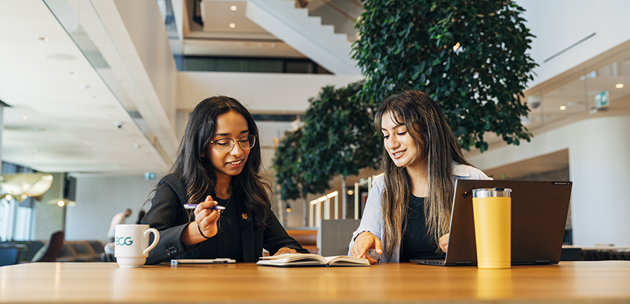 Two students sitting at a table in an open office space with a laptop, coffee mugs and a notebook reviewing notes