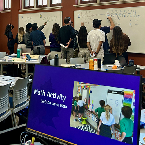 Student writing on white boards in a classroom with a computer monitor in the foreground reading "Math Activity"