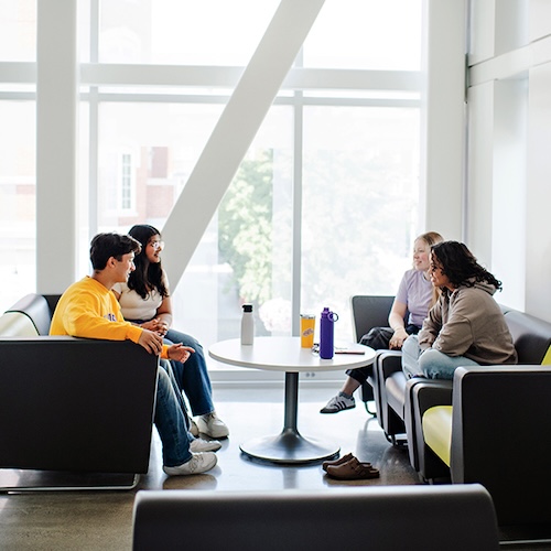 Four students sitting on couches in a lounge area with a large window in the background