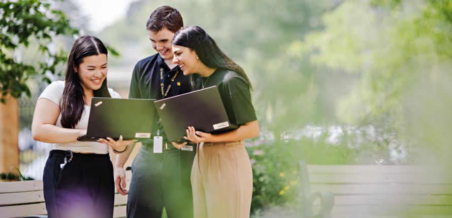 Three students standing in greenery looking at laptops.